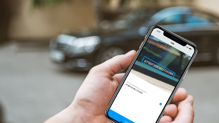 Hand in focus holding a phone with AI-powered VIN scanning technology on the screen with a car in the background